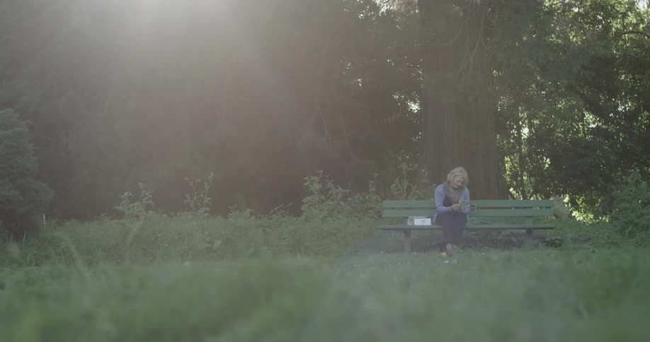 An old woman sits on a green bench in the sunlight