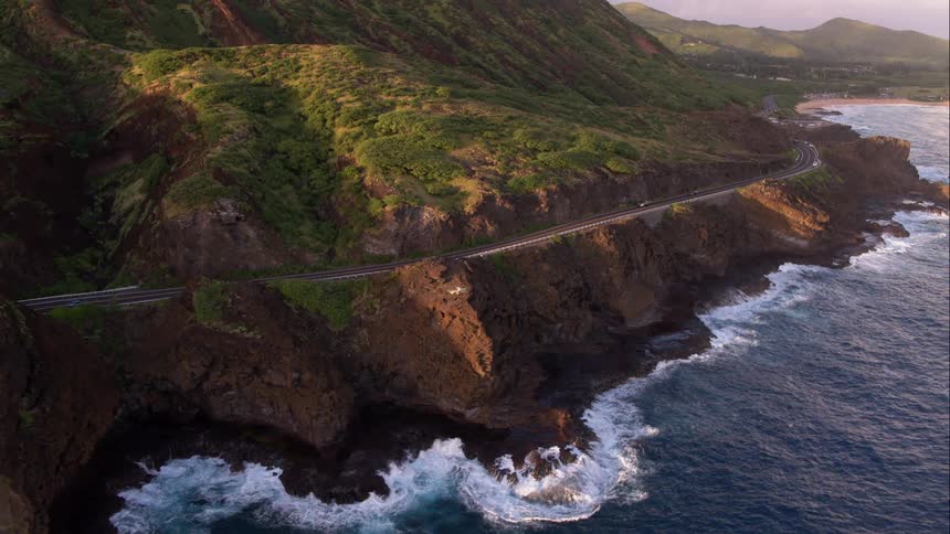 A road winds along the coast with waves crashing into the cliff below it