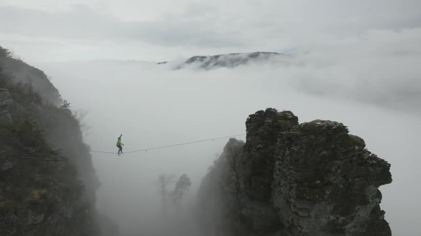 A person walks a tightrope between two mountain ridges with thick fog and clouds below