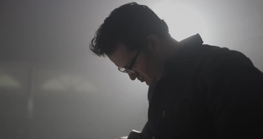 A man wearing glasses works on an electric guitar he is holding over a work table in his garage