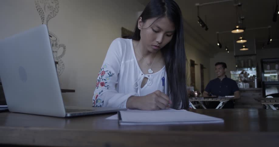 A woman looks at a laptop screen and writes something on a notepad