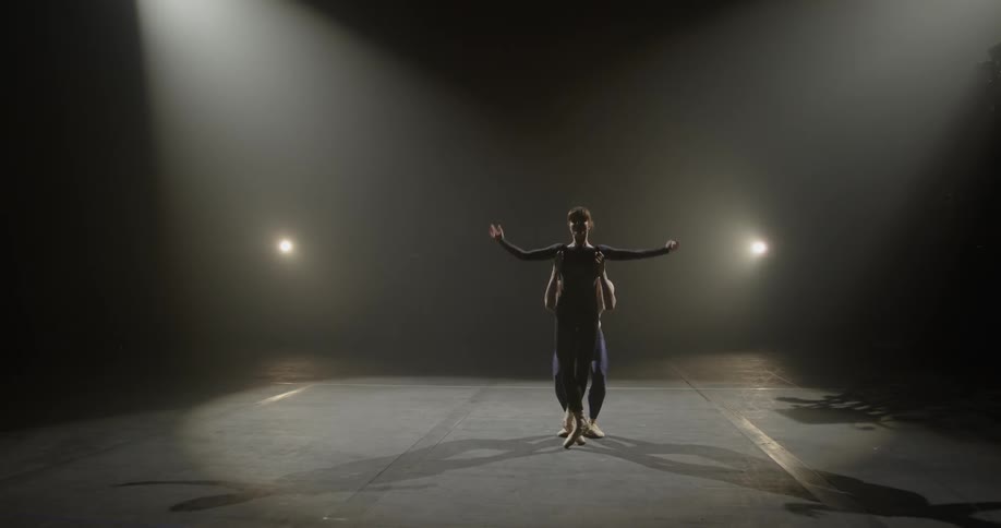 A man and a woman dance on a stage with two spotlights doing ballet dance routine in a dim lit room
