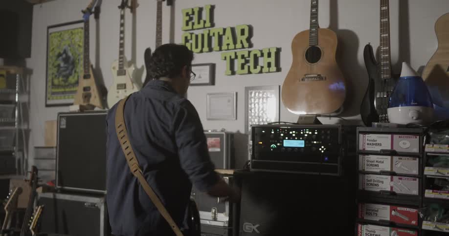 A man strums a guitar as he stands in front of several amps
