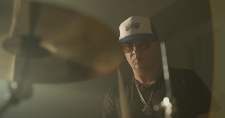 Drummer bangs on drum equipment wearing a hat and sunglasses in a dim lit room