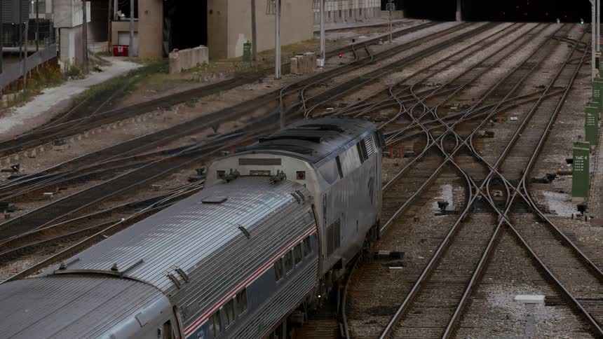 A train travels on a track with several switches heading towards a train station building