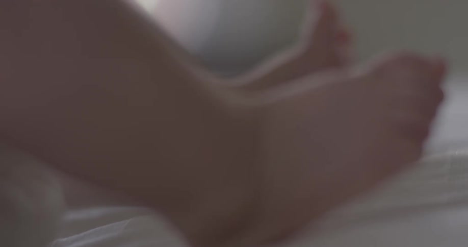 Baby feet are seen curling and moving around on a white bed sheet in a natural lighting room