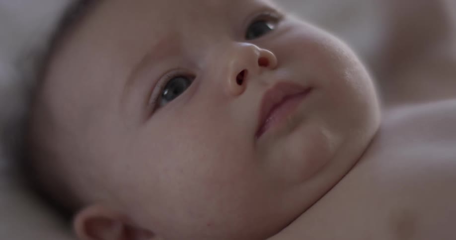 A baby looks towards the camera while laying down on a white blanket indoors and nearly cries