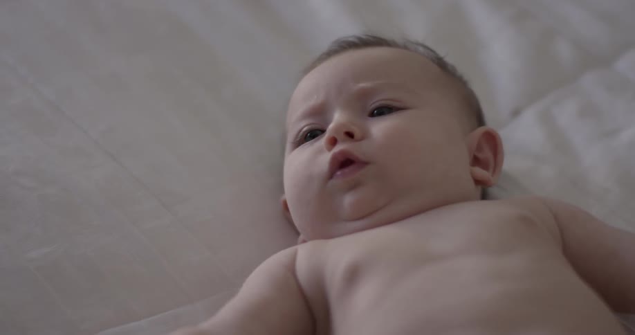 Baby reaches around trying to move and squirms with a pursed lip face laying on a blanket inside a room