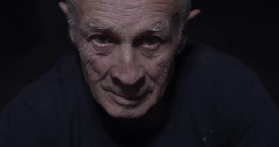 An old man sits in the darkness looking straight ahead