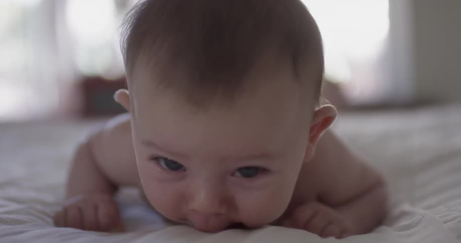 A baby lays face down on a white sheet as it tries to push itself up