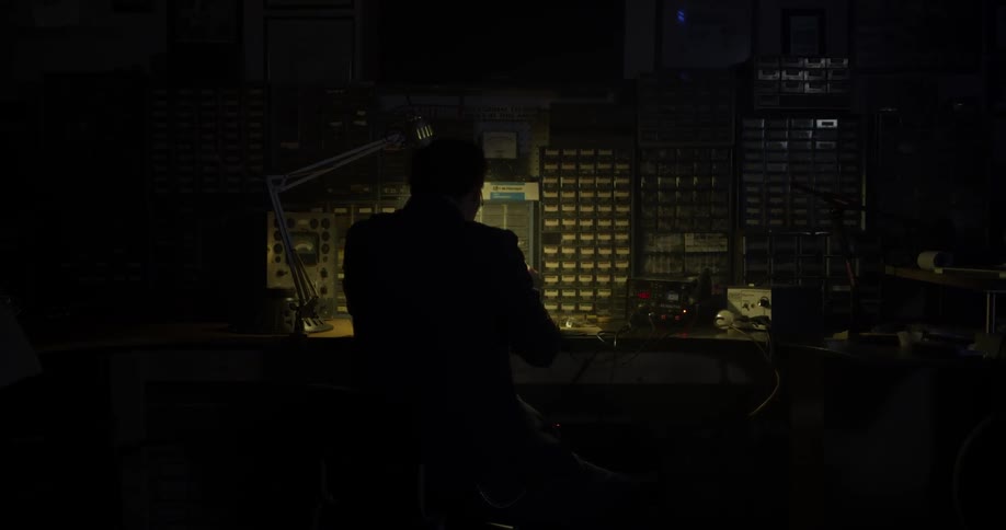 Someone sits in a dark workshop in front of a small light as a workbench