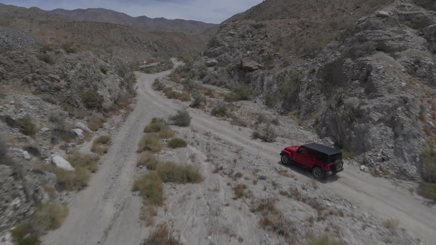 A car drives through the dirt road of a desert it is a red car and there are green shrubs around on a nice blue sunny day