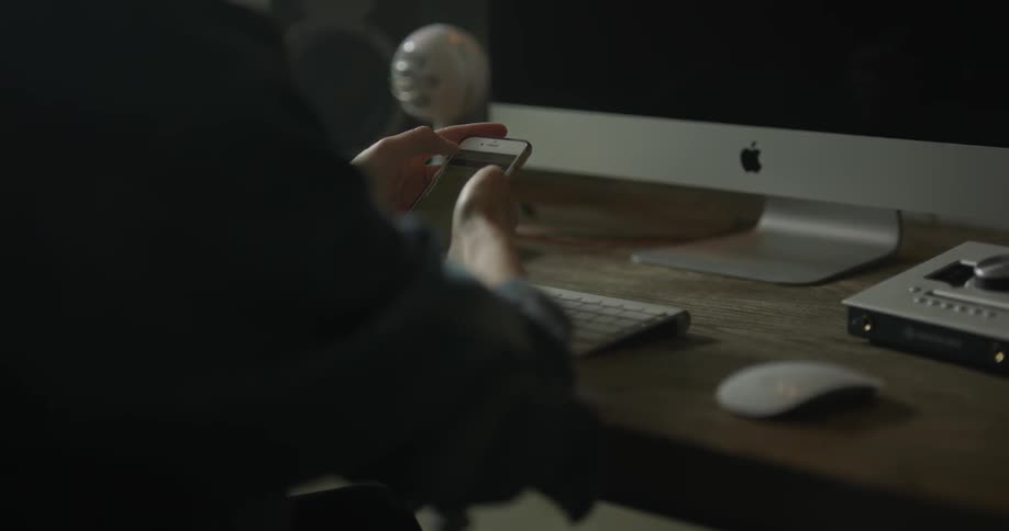 A woman sits at her wooden table desk holding her cellphone in front of a mac computer over a keyboard in a dimly lit room during the daytime