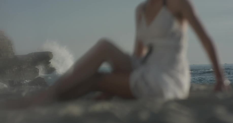 A woman in a white dress sits on the sand as waves splash up against the rocks