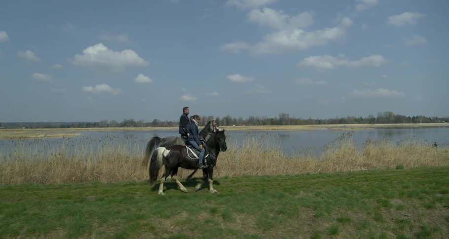 Two riders on horseback trot along a trail next to water