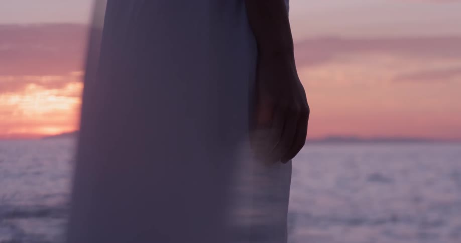 A woman stands on the beach in front of the ocean as the sun sets with red purple skies in a beautiful white dress