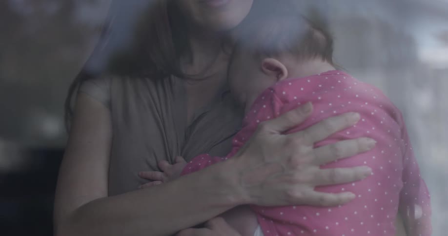 A woman holds a sleeping baby in front of a window