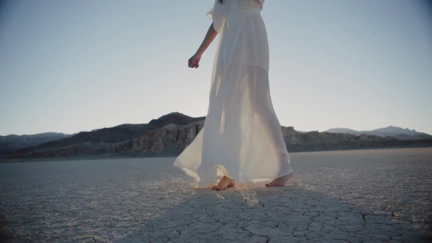 A woman wearing a white dress walking on dry dirt in the sun