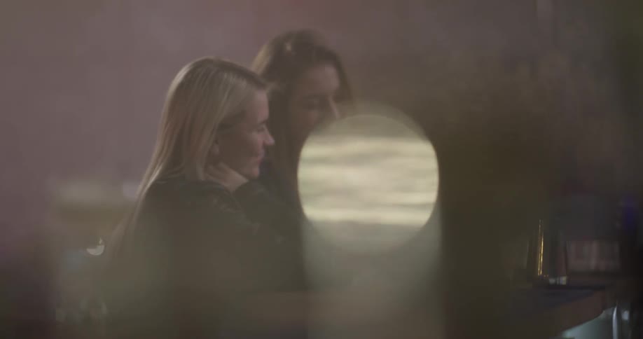 Two young women take selfies sitting at a bar