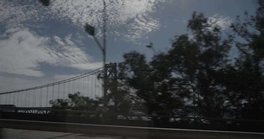 A car is driving by the underside of a bridge to see an opening to the blue skies full of clouds with a bridge and trees on the horizon