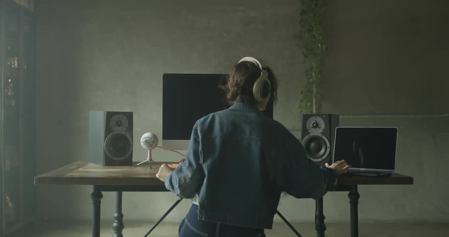 A woman is putting on headphones as she works at a desk with two computers