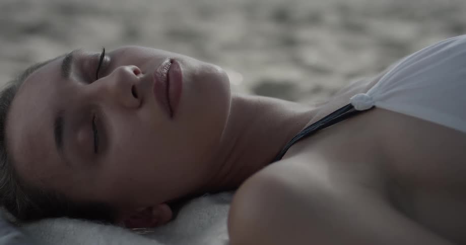 A woman is laying down in the sand with her eyes closes in the sunlight wearing a white dress