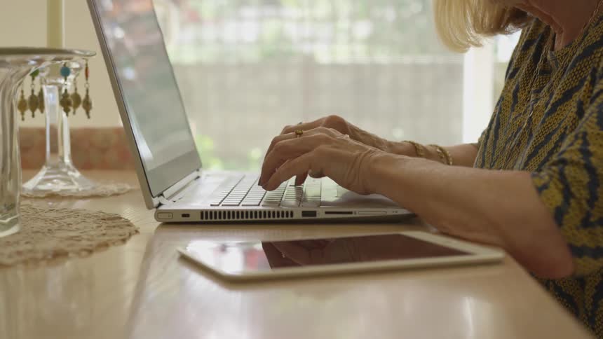 Woman types on laptop at table with tablet nearby
