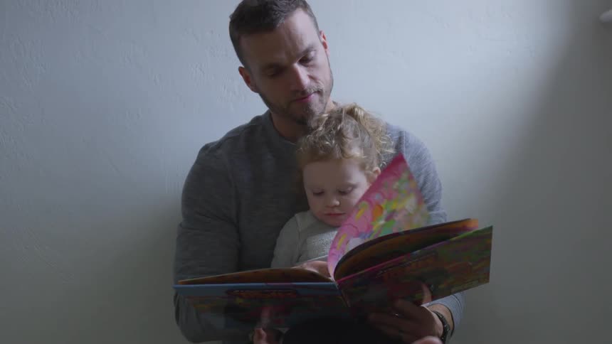 A man is sitting down against the wall with his girl child in his lap reading a book he is holding with sunlight coming in through the window above