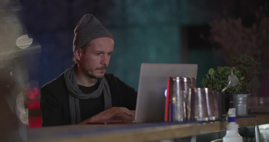 A man wearing a hat and scarf scrolls through the screen on his laptop