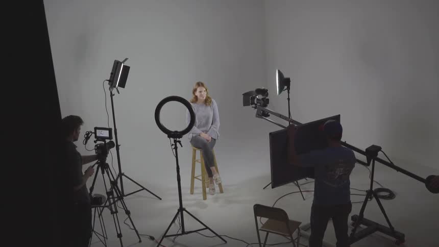 A woman sits in front of a white background with professional recording equipment pointed at her