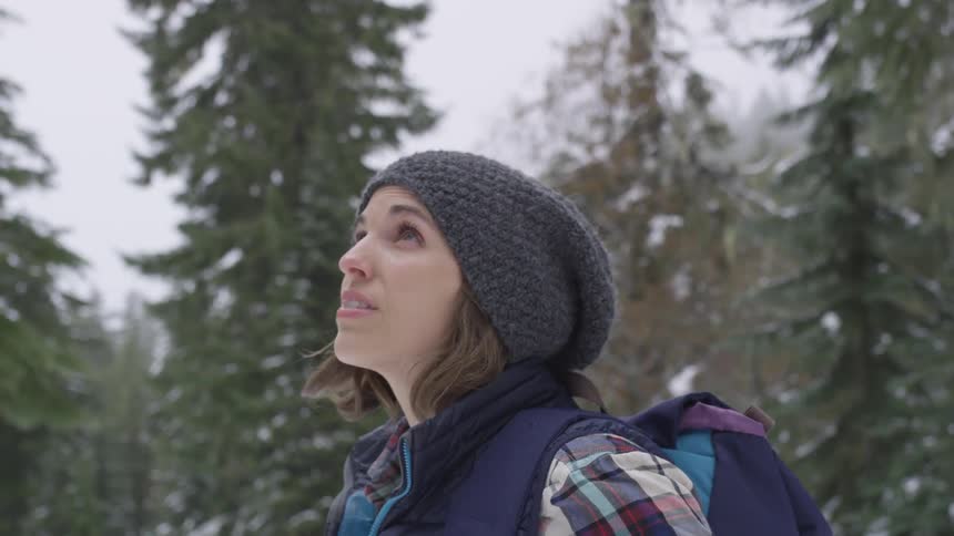 A woman stands looking up at a cloudy sky in the middle of a forest