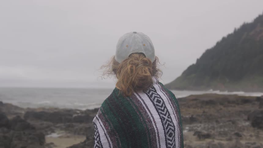 A girl with a cap on and a towel wrapped around her shoulders looks out at the ocean