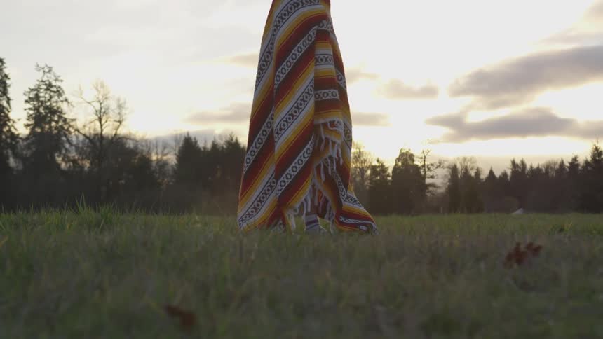 A person stands in a field wrapped in a blanket at dusk