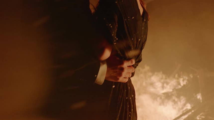 A man and a woman stand and hold each other by a fire