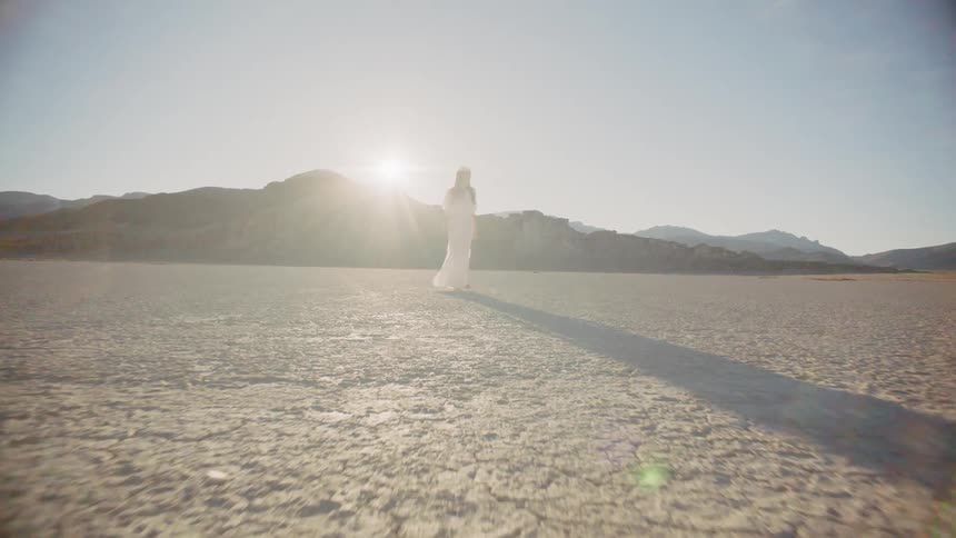 Zooming in to a woman standing in the middle of a desert with dry cracked soil rocks and mountains with a sunset and blue skies