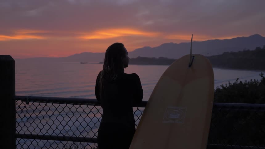 A woman stands next to a surfboard on a pier and looks out over the ocean at sunset