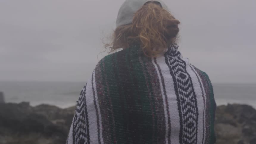 A woman wearing a hat and wrapped in a blanket stands on a rocky shore and looks out over the ocean