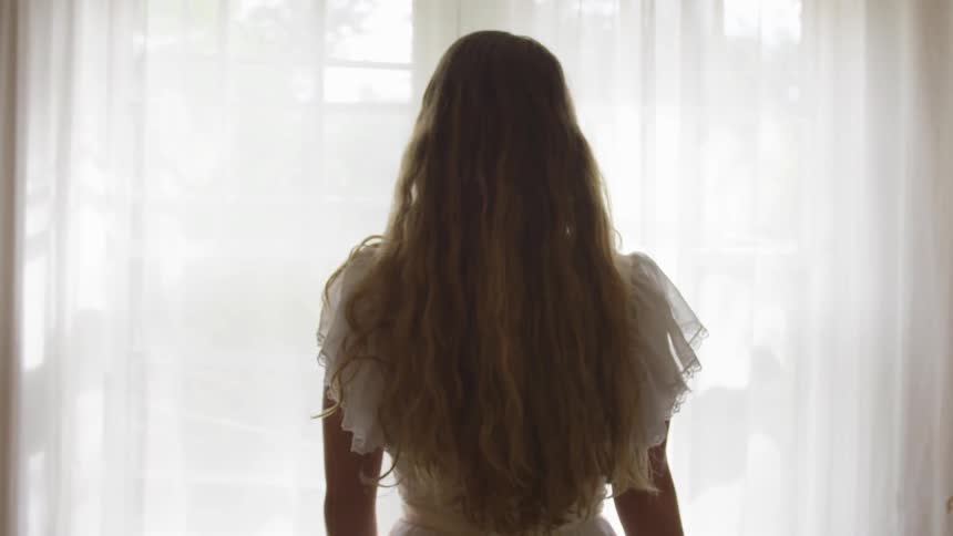 A woman standing in front of a window in a white dress