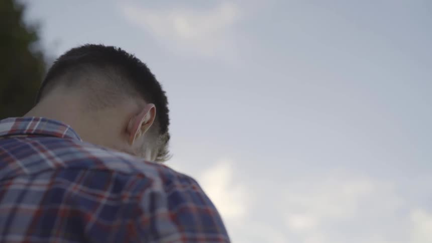 Man puts his head down and stands near green trees on a bright sunny day with some clouds