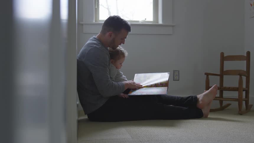 Man sitting on the floor reads a book to a toddler sitting on his lap