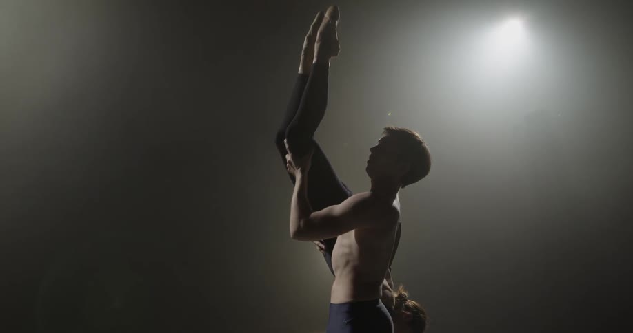 Man holding woman upside down while wearing tights in dark room with spotlight