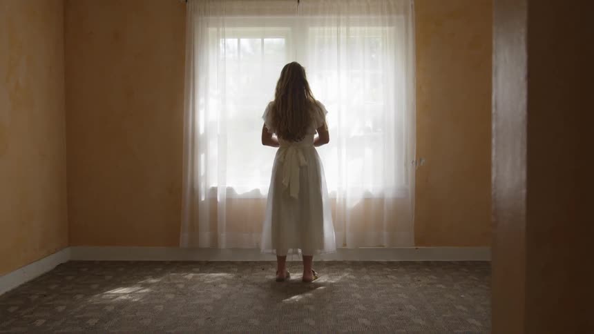 Blonde woman in a white dress standing in an empty room staring out a window