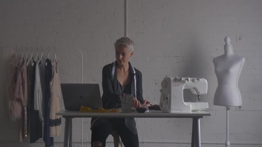 A woman opens her book and writes something down while staring at the laptop on her desk next to the sewing machine in a room for creating clothing