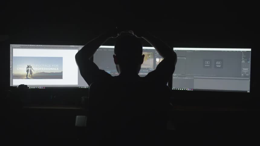 A man removes headphones and leans back in front of three monitors