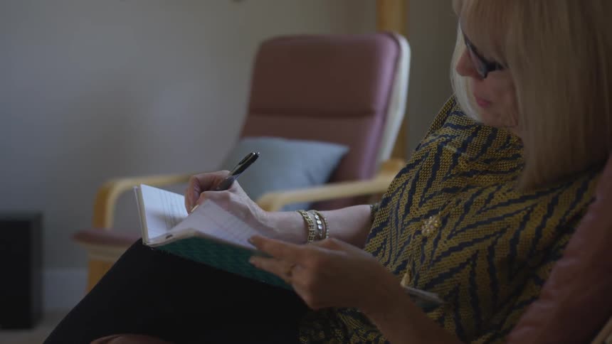 Elderly women sits on a chair and writes in a notebook with natural lighting from the window in her office
