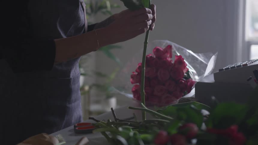 A woman holds flowers over a table adjusting and cutting petals trimming them to perfection to prepare for a bouquet in a dim lit room