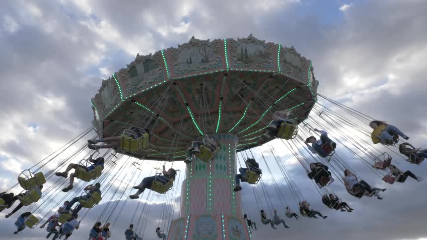 Many people sit in chairs on a ride at the amusement park as the camera looks up at the blue cloudy sky above on a bright sunny day