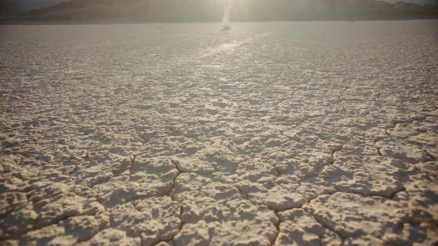 Cracked dry land in the desert as a lady in a white dress clothes wave in the background with the sun creating lens flares on a bright sunny day in the desert