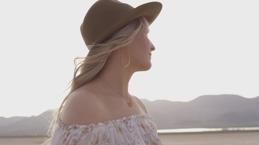 A woman stands in from of the sun in a valley of a mountain range on a bright sunny day wearing a hat blond hair and a white dress