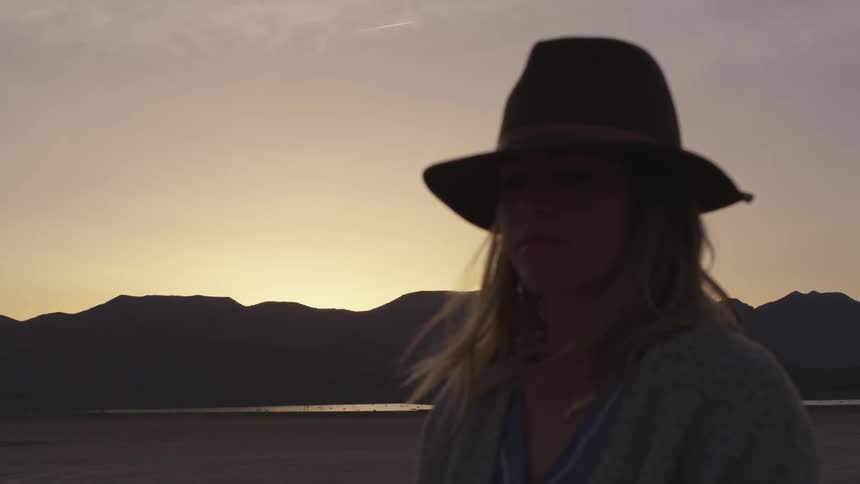 A woman wearing a hat stands outside near the mountains
